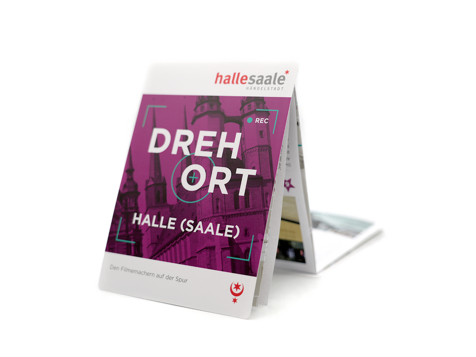 Der gefaltete Kulturstadtplan „Drehort Halle (Saale)“ steht auf weißem Untergrund. Sein violetter Einband mit weißer Schrift zeigt eine Stadtansicht, hebt Filmproduktionen hervor, präsentiert Drehorte und zeigt oben das Logo „halle saale“ mit rotem Stern.