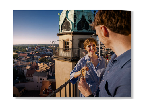 Eine Frau und ein Mann stehen bei Sonnenuntergang auf einem Dachbalkon und stoßen mit Champagner an, während sich das alte Gebäude mit der grünen Kuppel über Halle erhebt. Sie teilen ein Lächeln und erleben ein romantisches Abenteuer mit der Erlebnis-Box „Verliebt in Halle“ Classic.