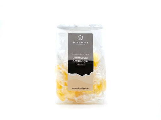 In einer durchsichtigen Plastiktüte sind einzeln verpackte gelbe Bonbons mit Orangen-Kurkuma-Geschmack zu sehen. Auf dem Etikett steht „Soole eG, Hallesche Schomgse Salzbonbons“, und die versiegelte Verpackung ist auf weißem Grund angebracht.