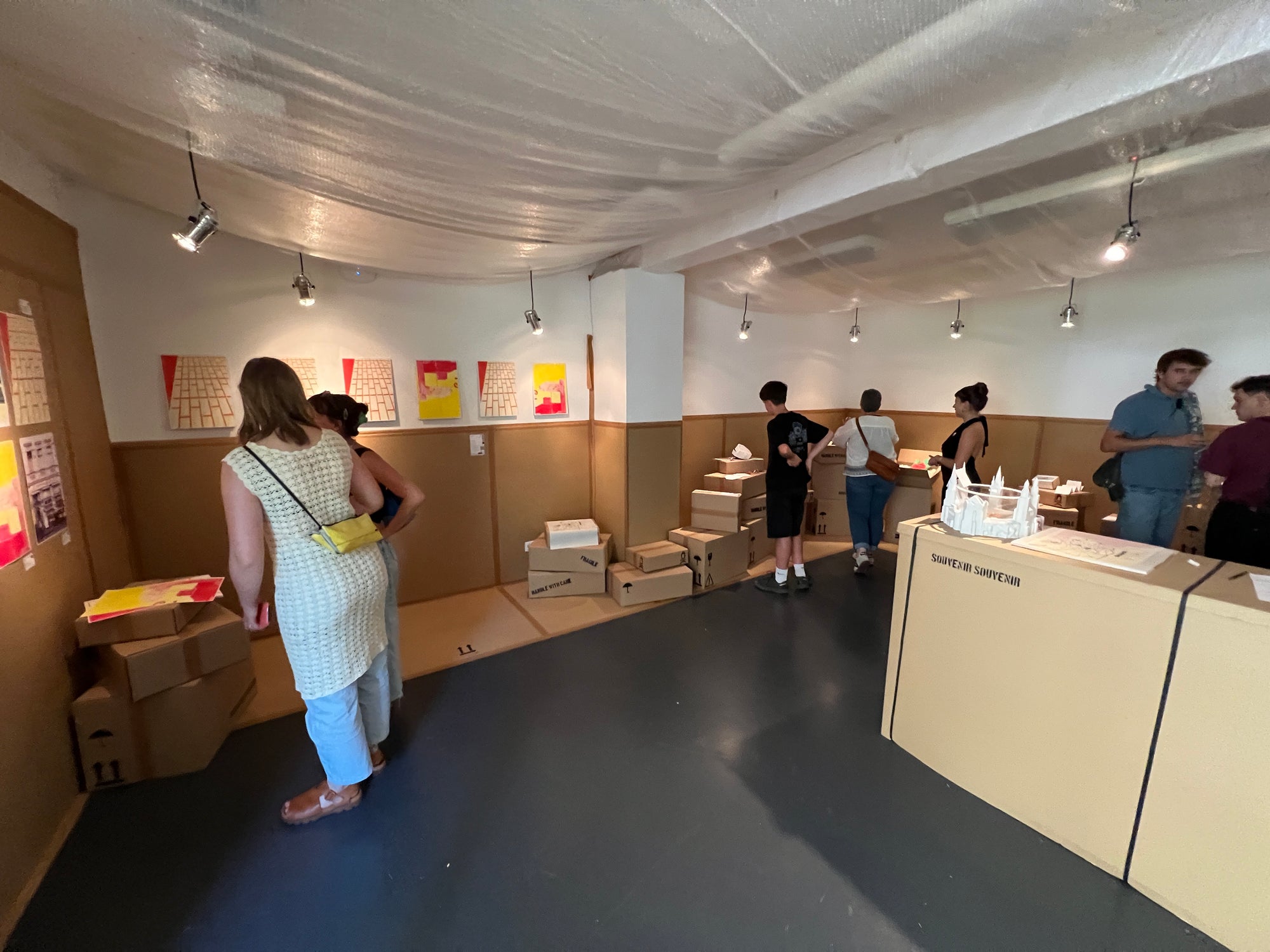Mehrere Personen erkunden einen kleinen Ausstellungsraum mit braunen Pappwänden und Ausstellungstischen. An den Wänden hängen Kunstwerke und Diagramme, auf den Tischen sind maßstabsgetreue Modelle und Objekte ausgestellt. Die Beleuchtung ist sanft, die Atmosphäre locker und einladend.