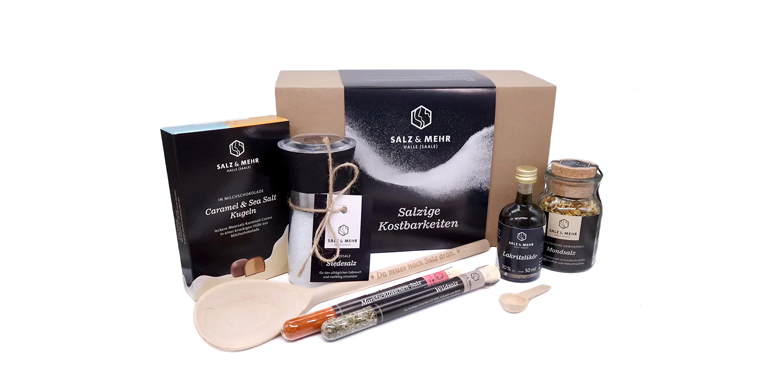 Ein Geschenkset mit Gourmet-Salzprodukten von Salz & Mehr umfasst ein Boxset, Reagenzgläser mit Gewürzen, einen Holzlöffel, ein Glas mit Kräutern, eine Flasche Öl, eine kleine Holzschaufel und eine Kerze, alles vor einem weißen Hintergrund arrangiert.