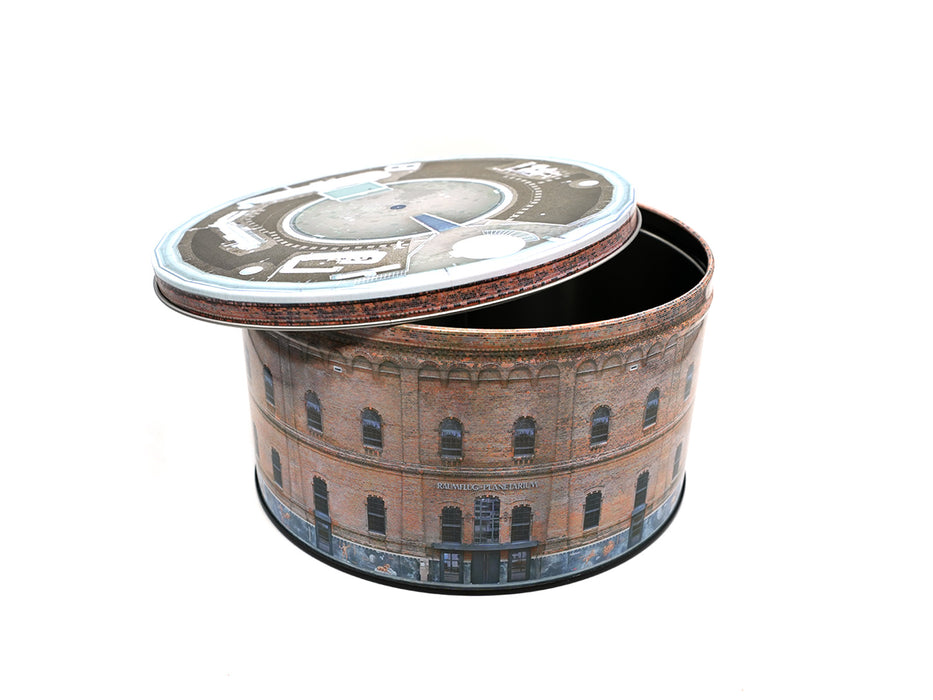 Die Keksdose „Planetarium“ ist eine runde Metalldose mit teilweise geöffnetem Deckel, die auf der Seite die Backsteinfassade des Planetariums Halle und auf dem Deckel eine Luftaufnahme des Daches zeigt, alles vor einem schlichten weißen Hintergrund.