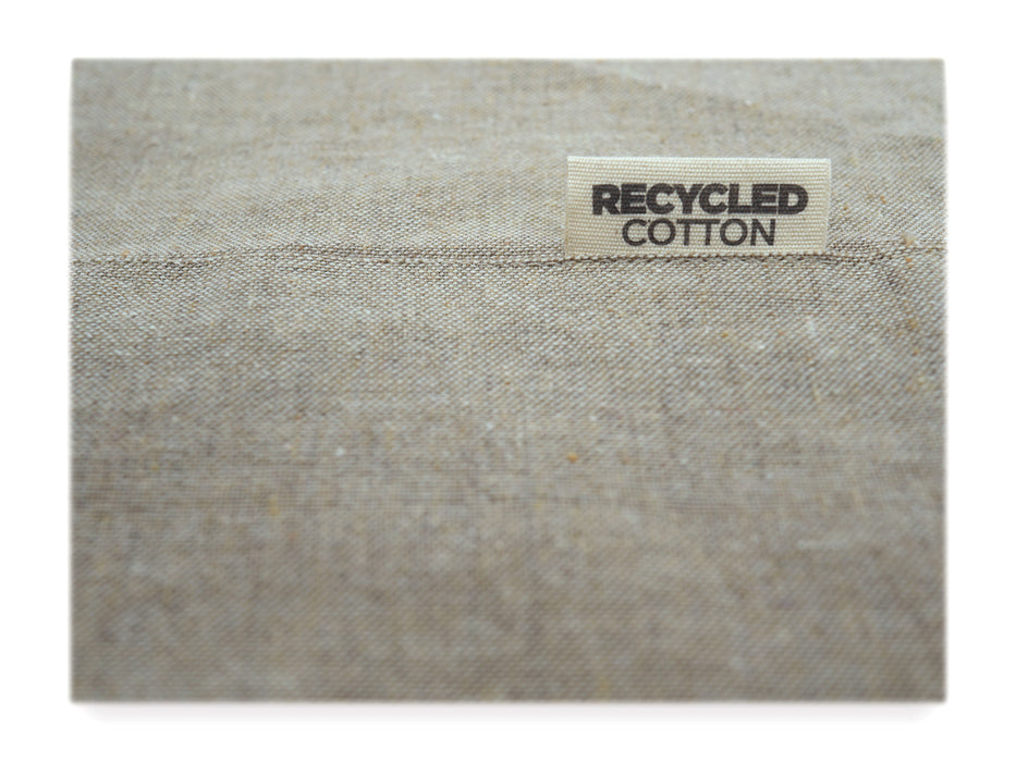 Eine Nahaufnahme von beigem Stoff mit gewebter Textur. Ein weißes Etikett mit der Aufschrift „RECYCLED COTTON“ hebt hervor, dass die Kochschürze „Versalzen? Nein, mit Liebe gekocht!“ aus recyceltem Material hergestellt ist.