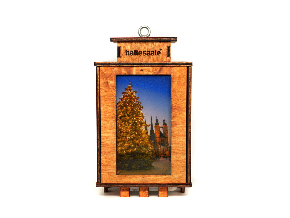 Die Holzlaterne „Halle (Saale)“ ist ein rechteckiges Holzornament mit der Gravur „Hallesaale“ auf der Oberseite und einem beleuchteten Weihnachtsbaumfoto. Sie besteht aus nachhaltigen Materialien, verfügt über eine Metallschlaufe zum Aufhängen und ein laternenähnliches Design.