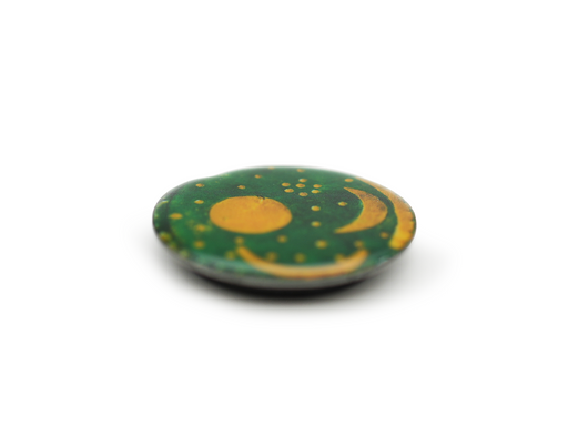 Der Magnet „Himmelsscheibe von Nebra“ besticht durch sein rundes, flaches Design mit dunkelgrüner Oberfläche und goldenen Motiven von Sonne, Halbmond und Sternen – eine Reminiszenz an die Himmelsscheibe. Präsentiert auf schlichtem, weißem Hintergrund wirken die Ränder sanft verschwommen.