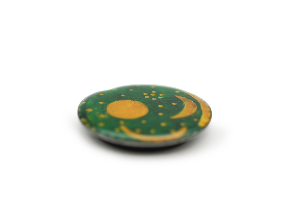 Der Magnet „Himmelsscheibe von Nebra“ besticht durch sein rundes, flaches Design mit dunkelgrüner Oberfläche und goldenen Motiven von Sonne, Halbmond und Sternen – eine Reminiszenz an die Himmelsscheibe. Präsentiert auf schlichtem, weißem Hintergrund wirken die Ränder sanft verschwommen.