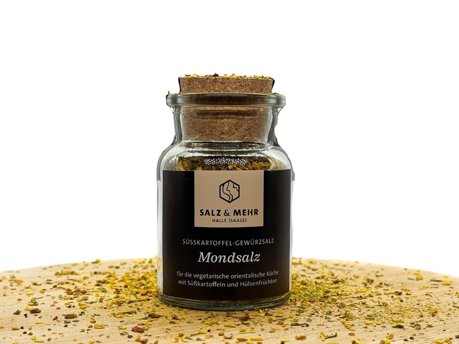 Ein kleines Glasgefäß mit der Aufschrift „Salz & Mehr, Mondsalz groß“ und Korkdeckel steht auf Holz, umgeben von lebendigen Gewürzen – ein ideales Gourmetsalz für die orientalische Küche.