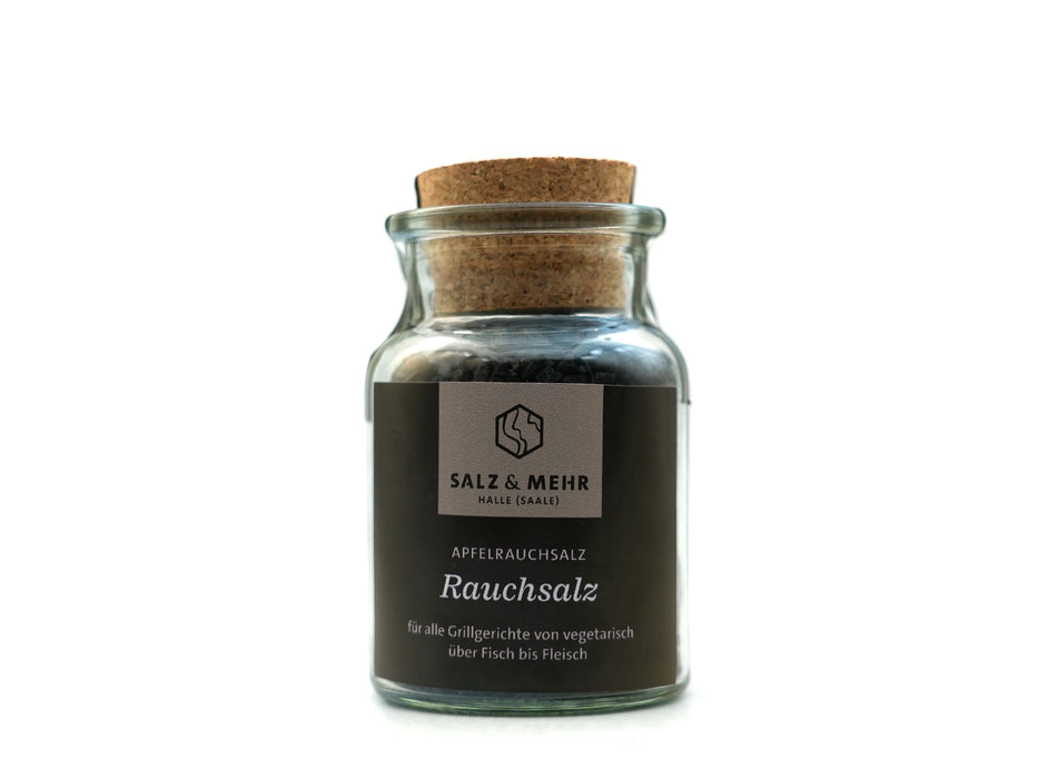 Ein klares Glasgefäß mit Korkdeckel enthält dunkles Apfelholz-Räuchersalz. Auf dem Etikett steht „Salz & Mehr-Bundle ‚Rauchsalz groß mit Salzschaufel‘“ und „für alle Grillgerichte von vegetarisch über Fisch bis Fleisch“. Weißer, übersichtlicher Hintergrund.