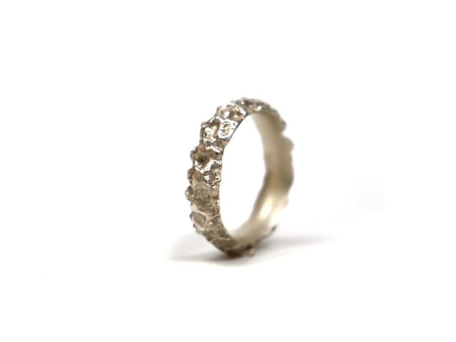 Der Ring „Saalesilber“ steht aufrecht auf einem schlichten weißen Hintergrund und präsentiert seine raue, natürliche, steinartige Textur und sein unregelmäßiges Band für ein unverwechselbares, organisches Aussehen anstelle einer glatten Oberfläche.