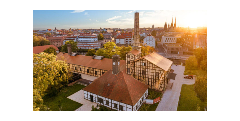 Luftaufnahme einer historischen europäischen Stadt bei Sonnenuntergang mit Fachwerkhäusern mit roten Ziegeldächern, einem zentralen Uhrenturm, von Bäumen gesäumten Straßen und einer Skyline mit Kirchtürmen und der fernen Stadtlandschaft unter einem klaren Himmel.