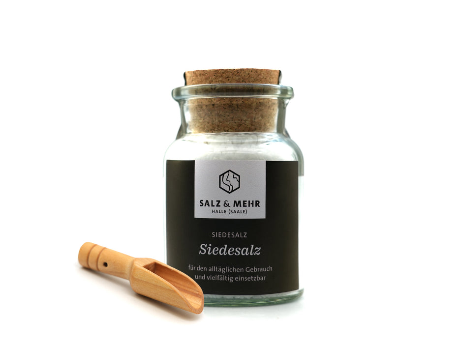 Das Siedesalz groß mit Salzschaufel von hallesaale* | Shop ist ein Korkenglas mit weißem Meersalz und Korkverschluss. Ein schwarzes Etikett mit weißem Text beschreibt es als ideal für den täglichen Gebrauch. Eine kleine Salzschaufel rundet das elegante Set ab.