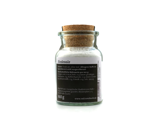 Ein durchsichtiges Glasgefäß mit der Aufschrift „Siedesalz groß“ (99,9 g) enthält weißes Speisesalz, ist mit einem Korken verschlossen und trägt ein deutsches Etikett mit der Aufschrift „Siedesalz, ohne Jod“, Nährwertangaben und dem Gewicht von 160 g. Das Gefäß steht vor einem weißen Hintergrund.