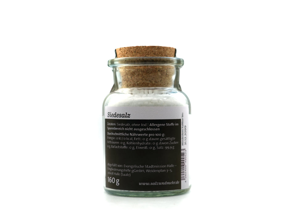 Ein durchsichtiges Glasgefäß mit der Aufschrift „Siedesalz groß“ (99,9 g) enthält weißes Speisesalz, ist mit einem Korken verschlossen und trägt ein deutsches Etikett mit der Aufschrift „Siedesalz, ohne Jod“, Nährwertangaben und dem Gewicht von 160 g. Das Gefäß steht vor einem weißen Hintergrund.