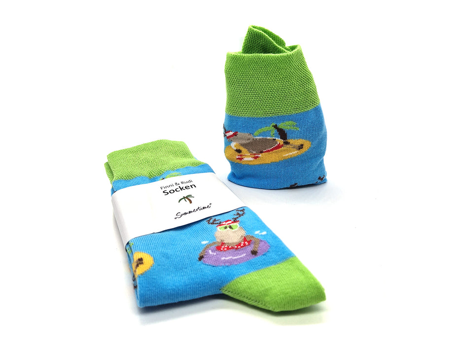 Das Socken-Set „Finni & Rudi“ Sommerzeit besteht aus zwei farbenfrohen Socken mit grünen Bündchen, Fersen und Zehen. Sie sind aus weicher Baumwolle gefertigt und nach OEKO-TEX Standard 100 zertifiziert. Sie zeigen ein blaues Stranddesign mit einem Hund mit Sonnenbrille, einer Palme und einer Sonne.