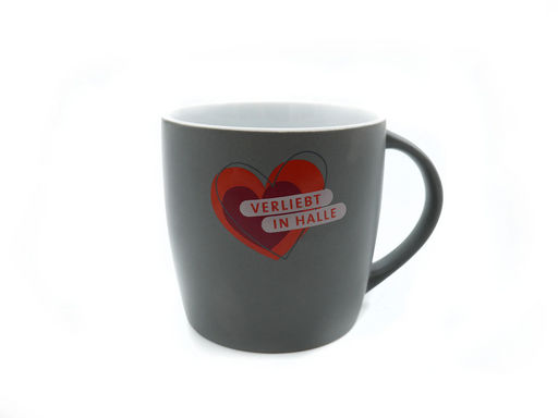 Eine dunkelgraue Tasse mit großem Henkel „Verliebt in Halle“ (grau-weiß) zeigt ein rotes Herz mit einem kleineren grauen Herzen und fettem weißen Text auf der Vorderseite: „VERLIEBT IN HALLE“. Die Tasse ist vor einem schlichten weißen Hintergrund abgebildet.