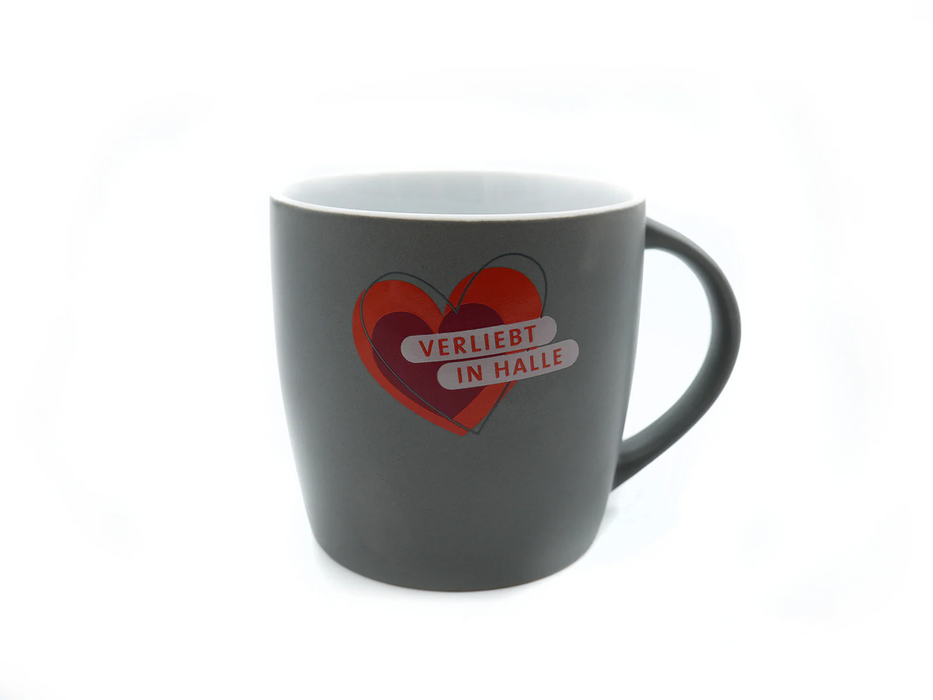 Eine dunkelgraue Tasse mit großem Henkel „Verliebt in Halle“ (grau-weiß) zeigt ein rotes Herz mit einem kleineren grauen Herzen und fettem weißen Text auf der Vorderseite: „VERLIEBT IN HALLE“. Die Tasse ist vor einem schlichten weißen Hintergrund abgebildet.