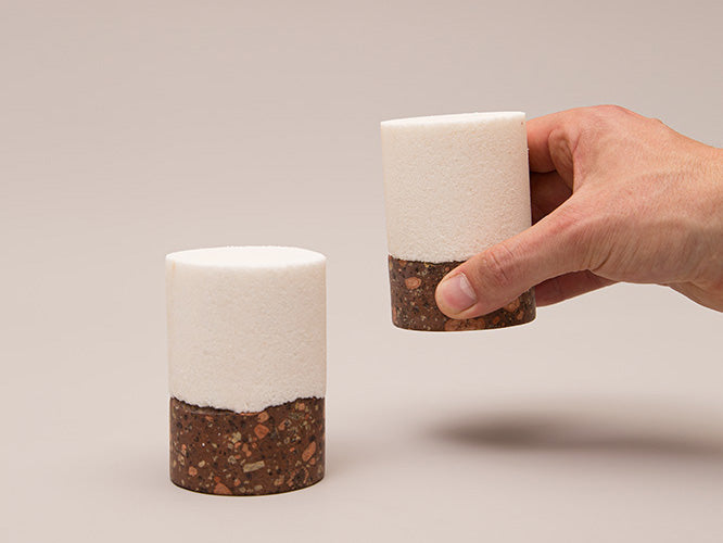 Eine Hand hält die Salzreibe „Pur“, eine kleine zylindrische Tasse mit weißem Deckel und strukturiertem braunem Löbejüner Porphyrboden. Eine weitere identische Tasse steht auf einer hellbeigen Fläche. Die kontrastierenden Materialien heben sich vom neutralen Hintergrund ab.