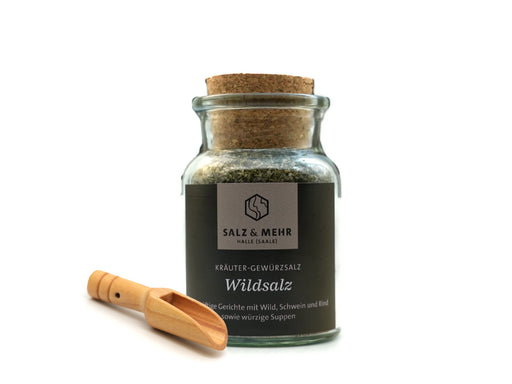 Ein Glasgefäß aus dem hallesaale* | Shop mit der Aufschrift „Salz & Mehr-Bundle: Wildsalz groß mit Salzschaufel“ trägt ein dunkles Etikett mit weißer Schrift und enthält eine Kräutersalzmischung. Eine kleine Holzschaufel auf weißem Grund begleitet das Gefäß.