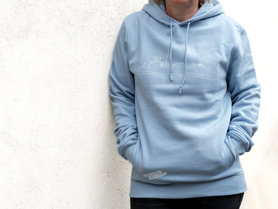 Eine Person steht vor einer weißen strukturierten Wand und trägt den „Verliebt in Halle“-Skyline-Halle-Hoodie mit einer minimalistischen Strichzeichnung auf der Brust. Ihre Hände stecken in den Taschen; schwarze Hosen und ein Teil ihres Gesichts sind teilweise sichtbar.