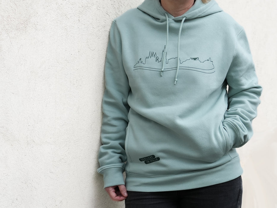 Eine Person trägt den hellblaugrünen Hoodie „Verliebt in Halle“ – Skyline Halle mit minimalistischer Skyline-Strichzeichnung auf der Brust, Kordelzügen, einem kleinen schwarzen Aufnäher am Saum und dem dezenten Text „Verliebt in Halle“ und steht vor einer weißen strukturierten Wand.