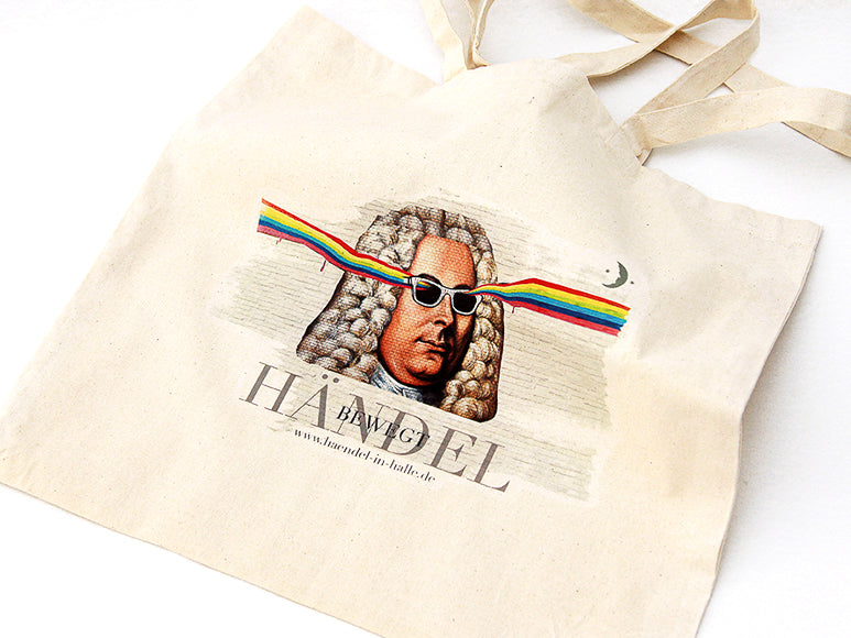Eine beige Tragetasche zeigt den Komponisten Händel mit Sonnenbrille, aus deren Gläsern bunte Regenbogenstrahlen schießen. Der Text darunter lautet „HÄNDEL“ mit darüberliegendem „BÄM G“ und der Webadresse „www.haendel-in-halle.de“.