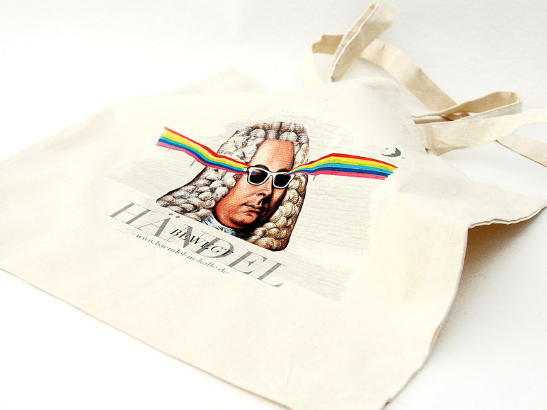 Der Leinenbeutel „Graffiti-Händel“ ist eine cremefarbene Fairtrade-Baumwolltasche mit der Abbildung eines Mannes mit gepuderter Perücke und weißer Sonnenbrille, der Regenbogenstrahlen aussendet. Unter dem Bild sind „HANDEL“ und ein kleinerer Text aufgedruckt.