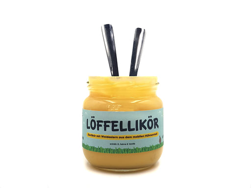Ein Glas mit der Aufschrift „MaMa‘s Unstruttaler Löffellikör“ (hellblaues Etikett mit schwarzer Schrift), gefüllt mit cremig-gelbem Eierlikör. Zwei Metalllöffel stehen aufrecht im Glas auf einer weißen Fläche vor weißem Hintergrund.