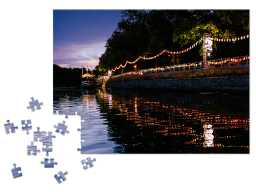 Ein teilweise fertiggestelltes Puzzle „Hier unten leuchten wir“ zeigt eine Flussszene in der Abenddämmerung während des Laternenfests mit einer Lichterkette, die sich im Wasser spiegelt, Bäumen am Ufer, einem violettblauen Himmel und verstreuten Teilen links vom Bild.