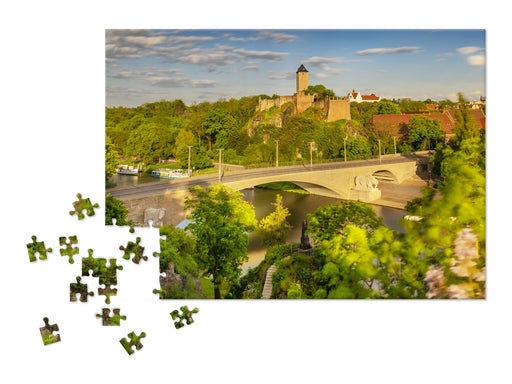 Ein teilweise fertiggestelltes Puzzle „Die Burg Giebichenstein in Halle (Saale)“ zeigt eine Steinbrücke über einen Fluss, grüne Bäume und eine historische Festung auf einem Hügel, wobei mehrere lose Teile auf einem weißen Hintergrund verstreut sind.