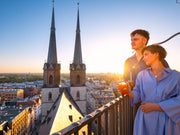 Eine Frau und ein Mann stehen bei Sonnenuntergang auf einem Dachbalkon und blicken mit einem Cocktail in der Hand über die Stadt. Sie erleben ein romantisches Abenteuer mit der Erlebnis-Box „Verliebt in Halle“ Piccolo.