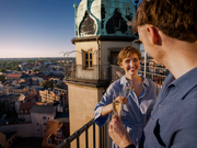 Eine Frau und ein Mann stehen bei Sonnenuntergang auf einem Dachbalkon und stoßen mit Champagner an, während sich das alte Gebäude mit der grünen Kuppel über Halle erhebt. Sie teilen ein Lächeln und erleben ein romantisches Abenteuer mit der Erlebnis-Box „Verliebt in Halle“ Classic.