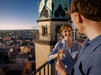 Eine Frau und ein Mann stehen bei Sonnenuntergang auf einem Dachbalkon und stoßen mit Champagner an, während sich das alte Gebäude mit der grünen Kuppel über Halle erhebt. Sie teilen ein Lächeln und erleben ein romantisches Abenteuer mit der Erlebnis-Box „Verliebt in Halle“ Classic.