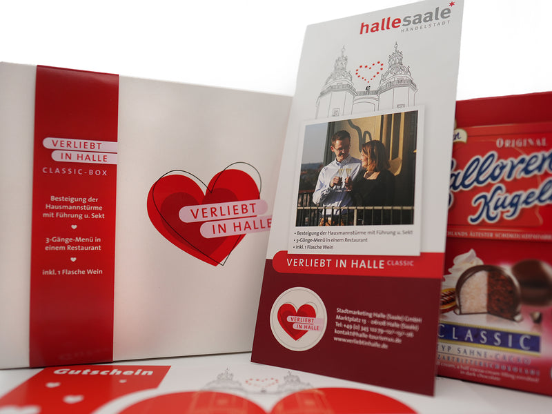 Eine Nahaufnahme der Erlebnis-Box „Verliebt in Halle“ Classic aus Halle (Saale), Deutschland, im Herz-Design mit weißer Box, rotem Herz, romantischer Broschüre für ein Candle-Light-Dinner für zwei und Halloren Kugeln Pralinen.