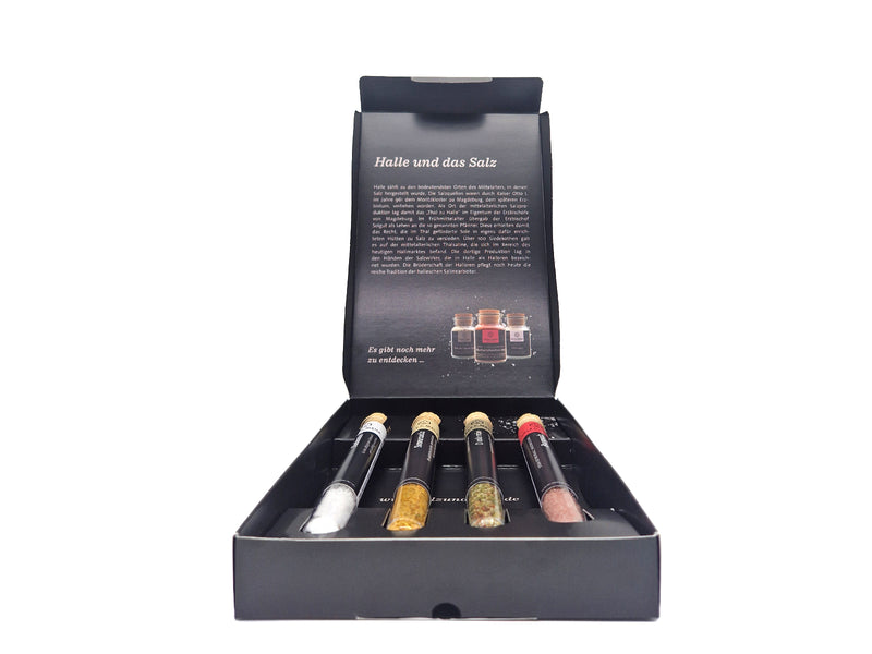 Geöffnete schwarze Geschenkbox „Kleine Salzschätze“ von SALZ & MEHR Halle (Saale) auf weißem Hintergrund. Im Deckel ist ein gedruckter Text über die Geschichte von „Halle und das Salz“ zu sehen, sowie Abbildungen von drei Gewürzgläsern. Im Bodenteil liegen in Einbuchtungen vier Reagenzgläser mit Korkverschluss, die mit unterschiedlichen Salzsorten und Gewürzmischungen gefüllt sind.