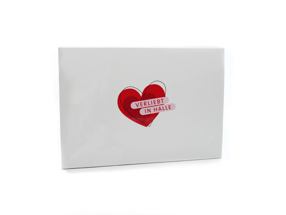Eine weiße, rechteckige Box mit einem roten Herz in der Mitte. Über dem Herzen befindet sich ein rosa Banner mit den roten Worten „Verliebt in Halle“. Der Hintergrund ist schlicht weiß.