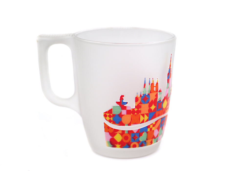 "Eine freigestellte, mattierte Milchglastasse mit einem quadratischen Henkel auf weißem Hintergrund. Der Text 'HALLE (SAALE)' ist in Rot darüber gedruckt. Darunter befindet sich ein farbenfrohes, geometrisches Mosaik, das die Skyline der Stadt Halle darstellt, zusammengesetzt aus bunten Quadraten, Kreisen und Dreiecken. Der untere Teil des Mosaiks ist durch ein unbedrucktes, bogenförmiges Glasband unterbrochen."
