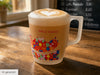 Nahaufnahme einer mattierten Glastasse mit Cappuccino, die auf einem Holztisch steht. Die Tasse ist mit einem roten Schriftzug 'HALLE (SAALE)' und einem bunten geometrischen Mosaikmuster, das eine Stadt-Silhouette darstellt, verziert. Der Milchschaum hat eine Herz-Latte-Art. Im unscharfen Hintergrund sind ein helles Fenster mit Pflanzen und eine dunkle Kreidetafel mit einem Menü und Preisen zu sehen. Unten rechts befindet sich ein kleines Label mit dem Text 'KI-generiert'.