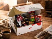 Eine weiß-goldene Geschenkbox mit der Aufschrift „Hallesche Genussbox“ steht geöffnet auf einem rustikalen Holztisch. Die Box ist mit Holzwolle gefüllt und enthält verschiedene Produkte wie Kaffee („Hallesche Röstung“), Halloren Kugeln, Fruchtsnacks und Likör. Im Vordergrund stehen eine gefüllte Kaffeetasse und eine Brille auf einem aufgeschlagenen Buch. Die Szene ist in warmes, gemütliches Licht getaucht.
