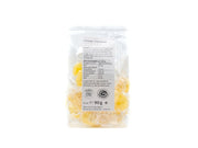 Eine transparente Plastiktüte mit Halleschen Schnongse-Salzbonbons „Orange-Curcuma“ ist aufrecht abgebildet, mit deutschen Nährwertangaben und Zutaten auf dem Etikett. Jedes gelbe Bonbon ist einzeln verpackt; Nettogewicht: 90 g.