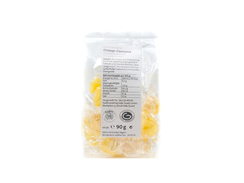 Eine transparente Plastiktüte mit Halleschen Schnongse-Salzbonbons „Orange-Curcuma“ ist aufrecht abgebildet, mit deutschen Nährwertangaben und Zutaten auf dem Etikett. Jedes gelbe Bonbon ist einzeln verpackt; Nettogewicht: 90 g.