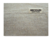 Eine Nahaufnahme von beigem Stoff mit gewebter Textur. Ein weißes Etikett mit der Aufschrift „RECYCLED COTTON“ hebt hervor, dass die Kochschürze „Versalzen? Nein, mit Liebe gekocht!“ aus recyceltem Material hergestellt ist.