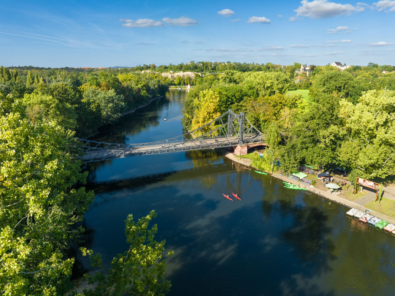Eine Metallbrücke überquert einen ruhigen, von Bäumen gesäumten Fluss unter blauem Himmel mit leichter Bewölkung. Zwei rote Kajaks paddeln darunter. Am Flussufer stapeln sich bunte Kajaks in der Nähe eines Stegs, umgeben von üppigem Grün. Im Hintergrund sind Häuser und Wald zu sehen.