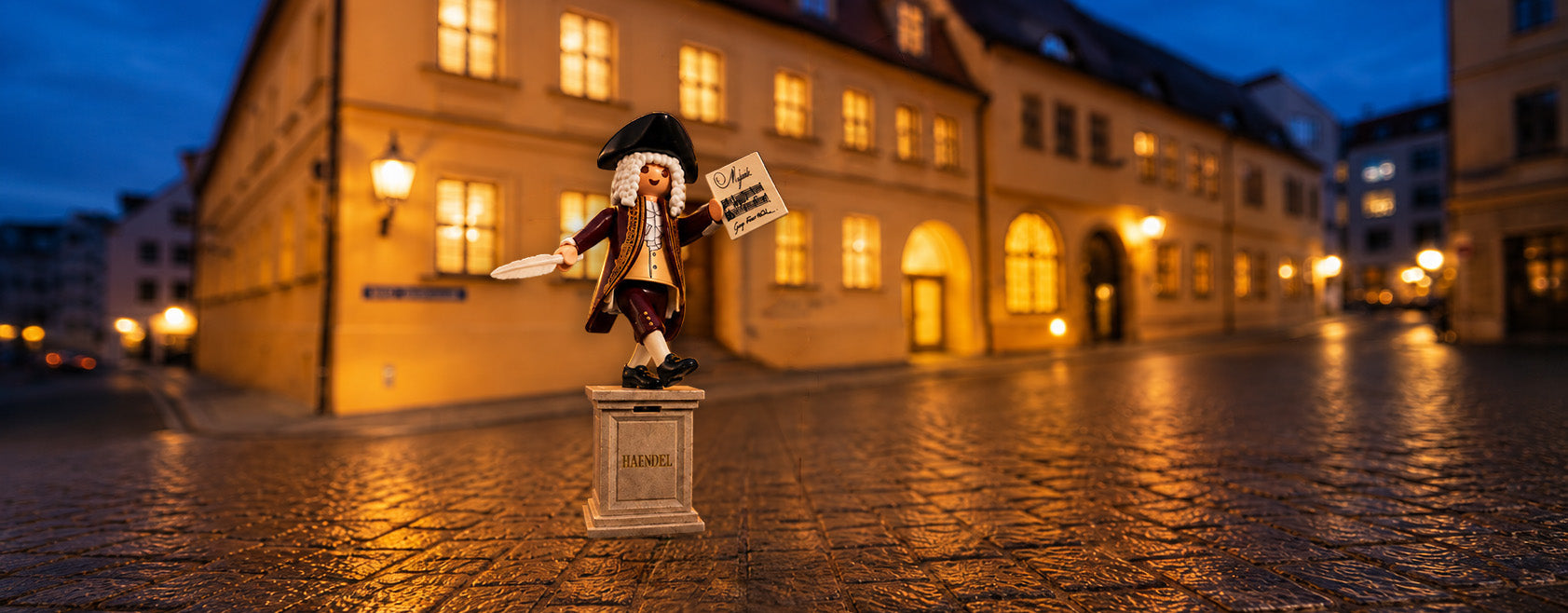 Eine Playmobil-Spielfigur des Komponisten Georg Friedrich Händel steht auf einem kleinen Sockel mit der Aufschrift „HAENDEL“ auf einem Kopfsteinpflaster. Die Figur trägt eine weiße Perücke, einen barocken Mantel und hält eine Schreibfeder sowie ein Notenblatt in den Händen. Im Hintergrund ist ein historisches, warm beleuchtetes Gebäude zur blauen Stunde zu sehen (Händel-Haus in Halle).