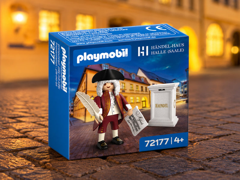 Die blaue Produktverpackung der Playmobil-Sonderfigur „Georg Friedrich Händel“ (Artikelnummer 72177) steht auf einem historischen Kopfsteinpflaster. Auf der Verpackung ist das Logo von Playmobil und das Logo des Händel-Hauses Halle (Saale) abgebildet. Das Verpackungsdesign zeigt die Händel-Figur mit Feder und Notenblatt vor dem beleuchteten Händel-Haus. Der Hintergrund des echten Bildes ist weichgezeichnet und zeigt eine warm beleuchtete Abendkulisse.