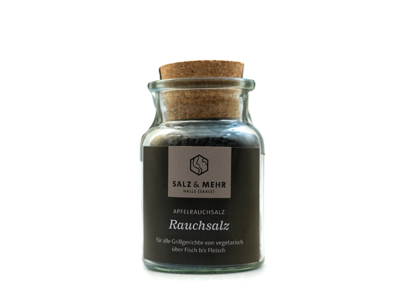 Ein klares Glasgefäß mit Korkdeckel enthält dunkles Apfelholz-Räuchersalz. Auf dem Etikett steht „Salz & Mehr-Bundle ‚Rauchsalz groß mit Salzschaufel‘“ und „für alle Grillgerichte von vegetarisch über Fisch bis Fleisch“. Weißer, übersichtlicher Hintergrund.