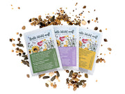 Drei bunte Samenpäckchen mit der Aufschrift „Samenmix ‚Bienenwiese‘“ und Blumen- und Bienenabbildungen liegen zwischen verstreuten Samen und Hülsen. Jedes Päckchen enthält eine einzigartige Wildblumenmischung zur Unterstützung der Bienen.