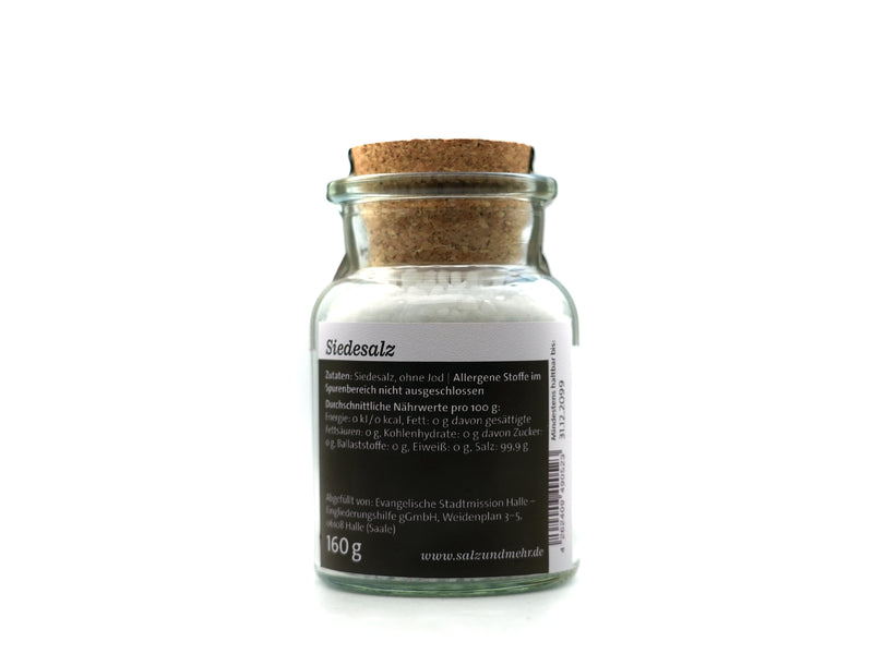 Ein durchsichtiges Glasgefäß mit der Aufschrift „Siedesalz groß“ (99,9 g) enthält weißes Speisesalz, ist mit einem Korken verschlossen und trägt ein deutsches Etikett mit der Aufschrift „Siedesalz, ohne Jod“, Nährwertangaben und dem Gewicht von 160 g. Das Gefäß steht vor einem weißen Hintergrund.