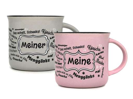 Produktfoto mit zwei Keramiktassen im Emaille-Becher-Stil vor weißem Hintergrund: Eine graue Tasse mit der Aufschrift „Meiner“ links hinten und eine rosa Tasse mit dem Schriftzug „Meine“ rechts im Vordergrund, jeweils umgeben von halleschen Dialekt-Wörtern.