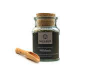 Ein Glasgefäß aus dem hallesaale* | Shop mit der Aufschrift „Salz & Mehr-Bundle: Wildsalz groß mit Salzschaufel“ trägt ein dunkles Etikett mit weißer Schrift und enthält eine Kräutersalzmischung. Eine kleine Holzschaufel auf weißem Grund begleitet das Gefäß.