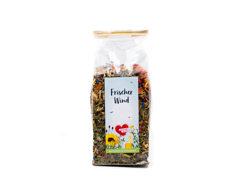 Ein durchsichtiger Plastikbeutel mit Tee „Frischer Wind“ trägt ein florales Etikett mit einem roten Herzen, das auf Minze- und Frühlingstee-Aromen hindeutet. Der Beutel, verschlossen mit einem goldenen Verschluss, ist auf schlichtem weißem Hintergrund abgebildet.